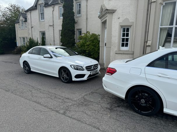 Two white cars parked along a narrow driveway beside a white building.
