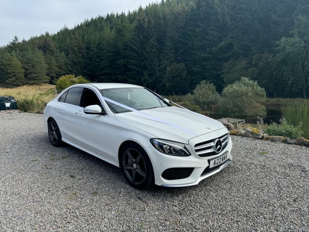 A white Mercedes-Benz C-Class parked on gravel with a forested hillside in the background.